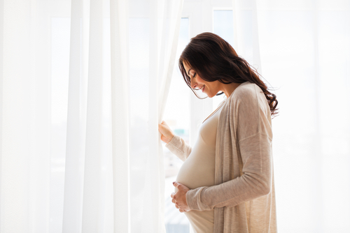 Picture of a smiling pregnant mother standing by a window holding a curtain open while she looks down at her stomach and holds it.