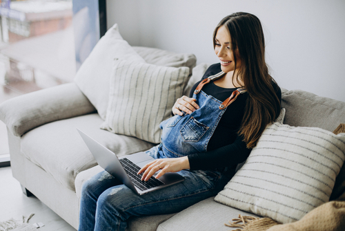 Picture of a pregnant mother sitting down on a couch holding her belly with one hand and the other hand is on a keyboard of an open laptop.