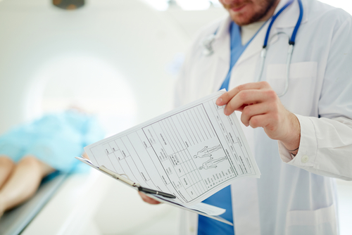 Picture of a male Physician wearing scrubs, a medical coat, and stethoscope around his neck. He is holding a clipboard while flipping through pages of  medical records.