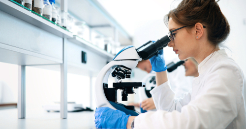 Picture of a female Pathologist wearing gloves and a medical coat. She is looking into a microscope.