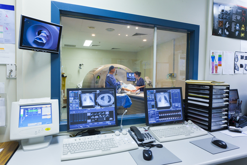 Picture of a Radiology Room showing a glass window, computers, and screens of MRI X-Rays. There is a MRI Machine that is shown in front of the window into another room. There is a male Radiologist and a female Nurse.
