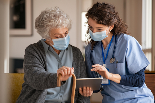Picture of a female Nurse sitting next to an elderly female patient. They are both wearing mask and looking down at a smart tablet.