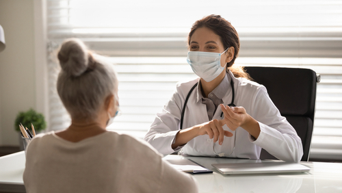 Picture of a female Physician sitting at a desk across from a female patient. They are both wearing mask.