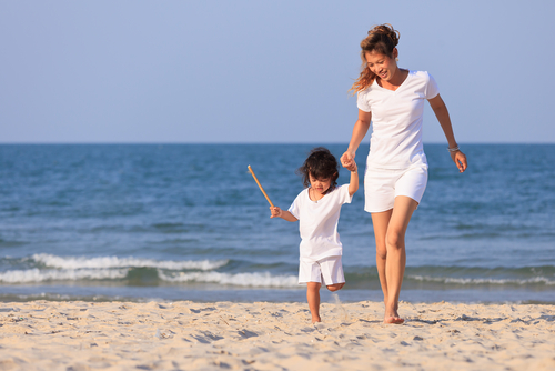 Picture of a mother and her toddler walking in front of a beach while holding hands and smiling. The toddler is holding a stick in one hand.