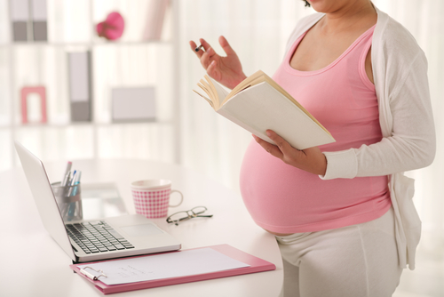Picture of a pregnant woman standing in front of a desk looking in a book and holding a pen in the other hand. There is a laptop, glasses, a clipboard, and cup of pens on the desk in front of her.