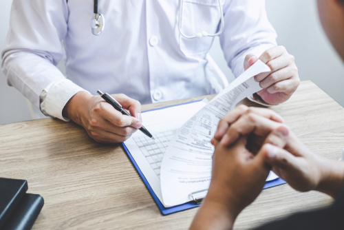 Picture of a male Physician sitting across from a male patient at a table. The Physician has a pen in one hand and clipboard with patient checklist paperwork in front of him.