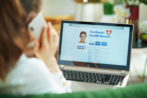 Picture of a female holding a smartphone up to her ear and she is sitting in front of an open laptop that says: Electronic Health Records (profile info).