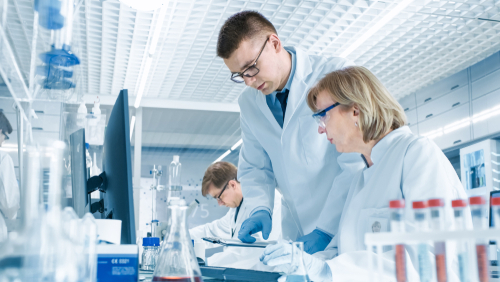 Picture of three lab techs (1 female and 2 males) in a Laboratory. They are wearing medical coats, medical glasses, and gloves looking down at paperwork on a clipboard.