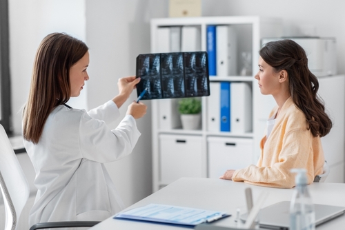 Picture of a female Physician holding up an X-ray while pointing  to it and looking at a female patient sitting down across from her.