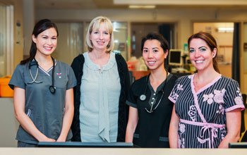 Picture of three smiling female Nurses and one smiling female Physician standing in the hospital hallway, close to The Nurses Station.