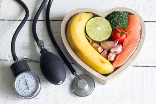 Picture of a blood pressure cuff, a stethoscope, and a wooden heart shape bowl that has a banana, lime, broccoli, garlic, tomato, and salmon in it.