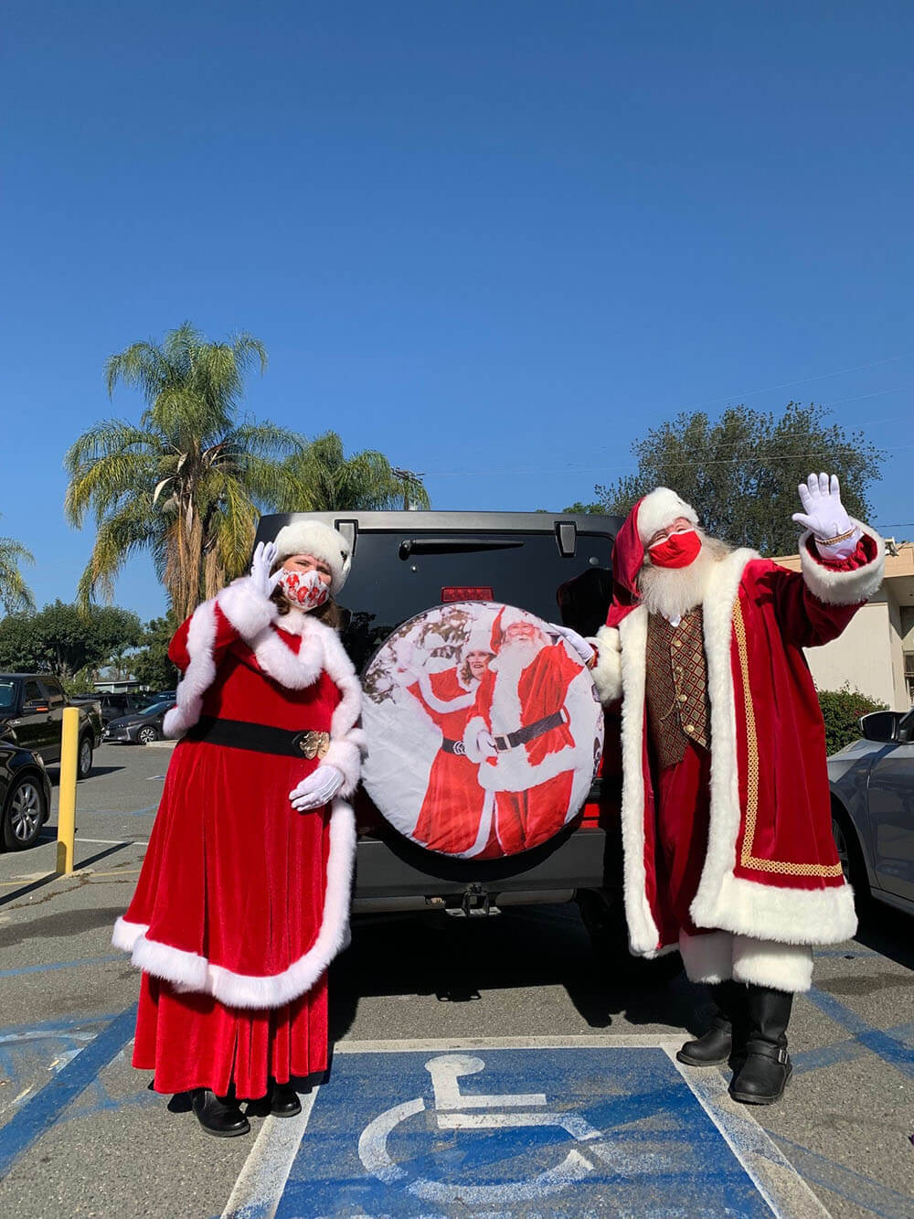 Santa and Mrs. Claus standing outside in the parking lot behind a Jeep that has a tire cover on the back of them